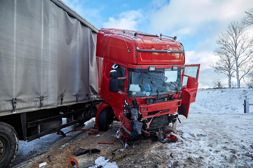 A red truck with a fabric-sided trailer is jack-knifed on a roadside lightly dusted with snow after a trucking accident.