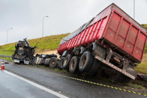 A red commercial truck is on the side of the road, tilted at an angle, with the cab of the truck separated from the rest. Driver fatigue may have contributed to this and other truck accidents.