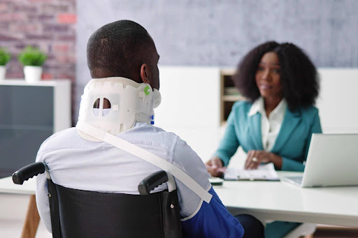 A man in a wheelchair wearing a neck brace and speaking to an attorney about medical expenses after a trucking accident.