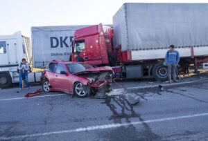 A small car, totaled after a truck accident, with three people observing the crash scene.