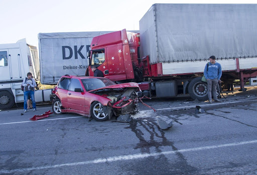 A small car, totaled after a truck accident, with three people observing the crash scene.